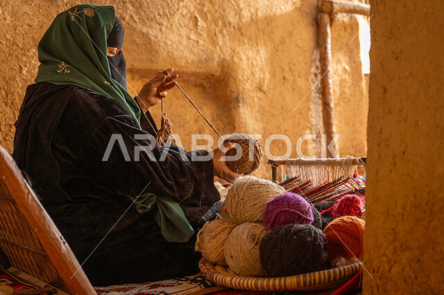 يوم بدينا 22 فبراير, المحافظة على التراث الشعبي السعودي, ذكرى تأسيس الدولة السعودية الأولى 1727م, حرفة يدوية تاريخيه, الشغف في ممارسة مهنة الغزل و النسج, صورة من الجانب لامرأة عربيه خليجية سعودية ترتدي البرقع و العباءه السوداء تقوم بلف خيوط الصوف