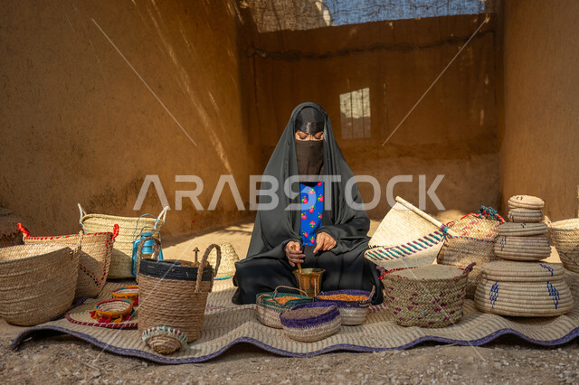 Preserving Saudi heritage, the day we started, February 22, the anniversary of the founding of the first Saudi state 1727 AD, displaying grains and local national products in baskets of hand-woven mats, a Saudi Arabian Gulf woman wearing a black abaya and burqa sitting on a popular rug grinding coffee beans in a copper grinder