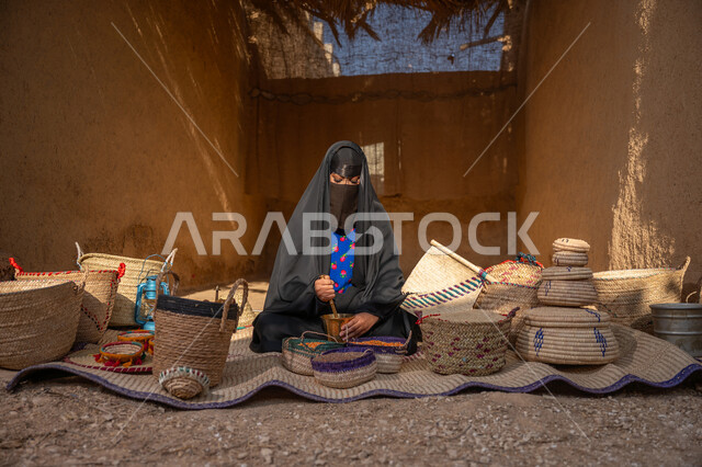 عرض الحبوب والمنتج المحلي الوطني في سلل من الحصير اليدوي، المحافظة على التراث السعودي، يوم بدينا 22 فبراير, ذكرى تأسيس الدولة السعودية الأولى 1727م, امرأة عربيه خليجية سعودية ترتدي العباءه السوداء و البرقع تجلس على بساط شعبي تقوم بطحن حبوب البن في النجر النحاسي