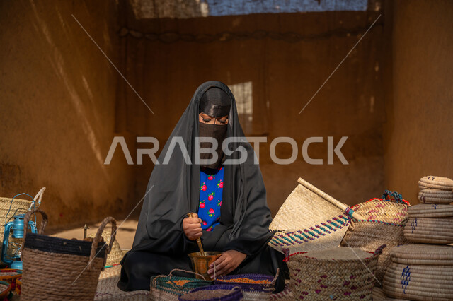 Anniversary of the founding of the first Saudi state 1727 AD, displaying grains and local national products in baskets of handmade mats, preserving Saudi heritage, the day we started, February 22, a Saudi Gulf Arab woman wearing a black abaya and burqa sitting on a popular rug grinding coffee beans in a copper grinder