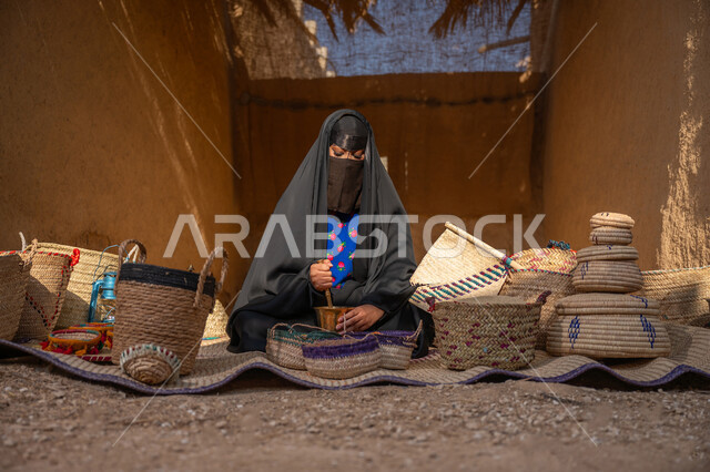 يوم بدينا 22 فبراير, ذكرى تأسيس الدولة السعودية الأولى 1727م, عرض الحبوب والمنتج المحلي الوطني في سلل من الحصير اليدوي، المحافظة على التراث السعودي، امرأة عربيه خليجية سعودية ترتدي العباءه السوداء و البرقع تجلس على بساط شعبي تقوم بطحن حبوب البن في النجر النحاسي