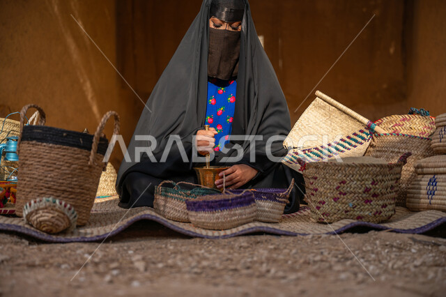 المحافظة على التراث السعودي، يوم بدينا 22 فبراير, ذكرى تأسيس الدولة السعودية الأولى 1727م, عرض الحبوب والمنتج المحلي الوطني في سلل من الحصير اليدوي، امرأة عربيه خليجية سعودية ترتدي العباءه السوداء و البرقع تجلس على بساط شعبي تقوم بطحن حبوب البن في النجر النحاسي