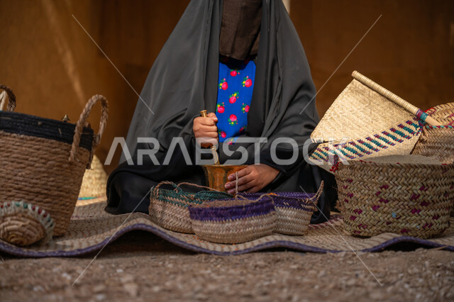 يوم بدينا 22 فبراير, ذكرى تأسيس الدولة السعودية الأولى 1727م, عرض الحبوب والمنتج المحلي الوطني في سلل من الحصير اليدوي، المحافظة على التراث السعودي، امرأة عربيه خليجية سعودية ترتدي العباءه السوداء و البرقع تجلس على بساط شعبي تقوم بطحن حبوب البن في النجر النحاسي