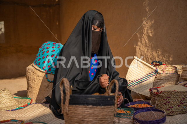 ذكرى تأسيس الدولة السعودية الأولى 1727م, عرض الحبوب والمنتج المحلي الوطني في سلل من الحصير اليدوي، المحافظة على التراث السعودي، امرأة عربيه خليجية سعودية ترتدي العباءه السوداء و البرقع تجلس على بساط شعبي تقوم بطحن حبوب البن في النجر النحاسي، يوم بدينا 22 فبراير