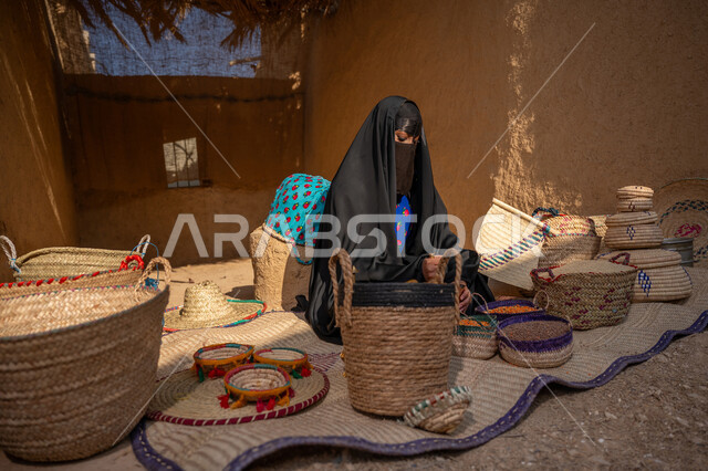 عرض الحبوب والمنتج المحلي الوطني في سلل من الحصير اليدوي، المحافظة على التراث السعودي، ذكرى تأسيس الدولة السعودية الأولى 1727م, امرأة عربيه خليجية سعودية ترتدي العباءه السوداء و البرقع تجلس على بساط شعبي تقوم بطحن حبوب البن في النجر النحاسي، يوم بدينا 22 فبراير