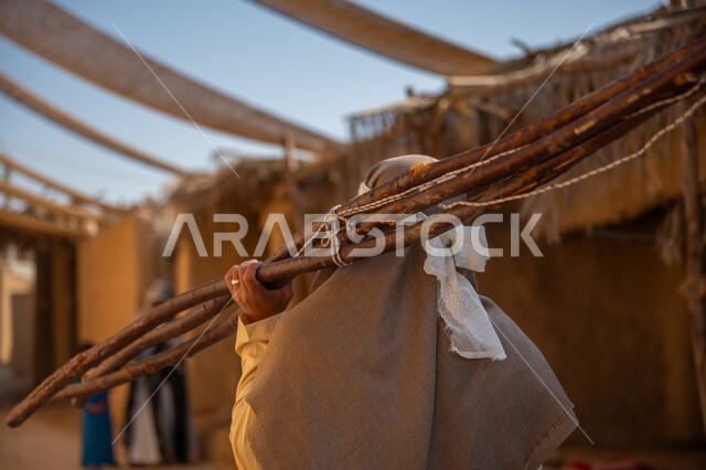 وسائل التدفئه القديمة، الحياة التقليدية البدوية، ذكرى تأسيس الدولة السعودية الأولى 22 فبراير، تجميع الحطب في موسم الشتاء, صورة مقربة من الخلف لفلاح عربي خليجي سعودي يحمل على كتفه مجموعه من أعواد الخشب الثقيلة داخل بيت طيني تراثي، يوم بدينا 1727 م