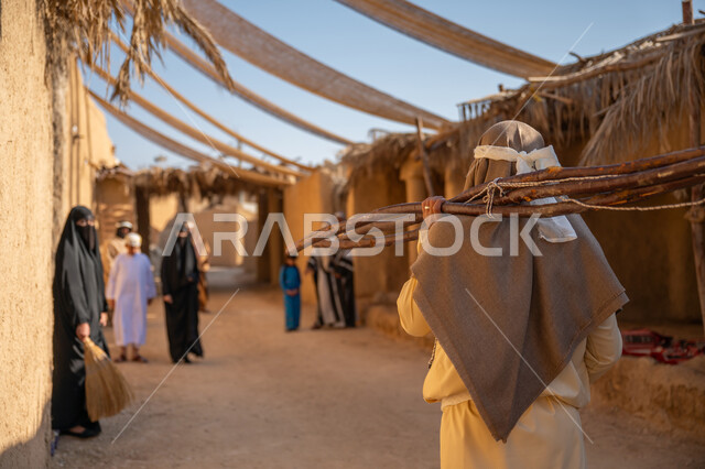 الحياة التقليدية البدوية، استخدام وسائل التدفئه القديمة، ذكرى تأسيس الدولة السعودية الأولى1727 م، يوم بدينا 22 فبراير، تجميع الحطب بموسم الشتاء, صورة مقربة من الخلف لرجل عربي خليجي سعودي يرتدي الثوب التقليدي والغترة يحمل على كتفه الخشب الثقيل ويمشي في حي تاريخي