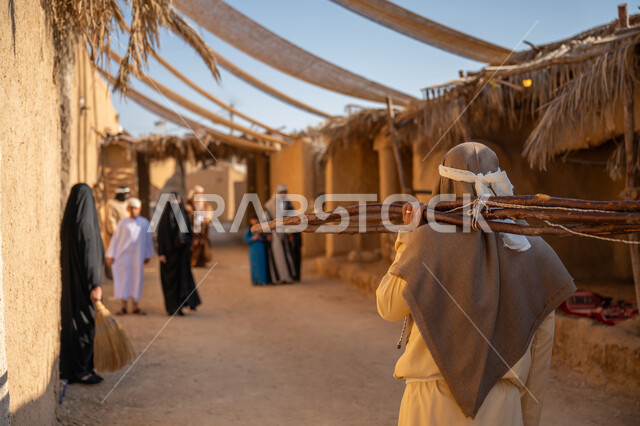 ذكرى تأسيس الدولة السعودية الأولى1727 م، الحياة التقليدية البدوية، استخدام وسائل التدفئه القديمة، يوم بدينا 22 فبراير، تجميع الحطب بموسم الشتاء, صورة مقربة من الخلف لرجل عربي خليجي سعودي يرتدي الثوب التقليدي والغترة يحمل على كتفه الخشب الثقيل ويمشي في حي تاريخي