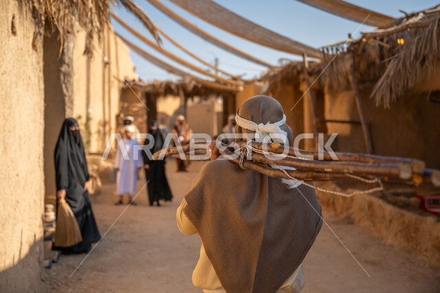 استخدام وسائل التدفئه القديمة، الحياة التقليدية البدوية، ذكرى تأسيس الدولة السعودية الأولى1727 م، يوم بدينا 22 فبراير، تجميع الحطب بموسم الشتاء, صورة مقربة من الخلف لرجل عربي خليجي سعودي يرتدي الثوب التقليدي والغترة يحمل على كتفه الخشب الثقيل ويمشي في حي تاريخي