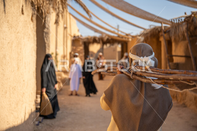 استخدام وسائل التدفئه القديمة، الحياة التقليدية البدوية، ذكرى تأسيس الدولة السعودية الأولى1727 م، يوم بدينا 22 فبراير، تجميع الحطب بموسم الشتاء, صورة مقربة من الخلف لرجل عربي خليجي سعودي يرتدي الثوب التقليدي والغترة يحمل على كتفه الخشب الثقيل ويمشي في حي تاريخي