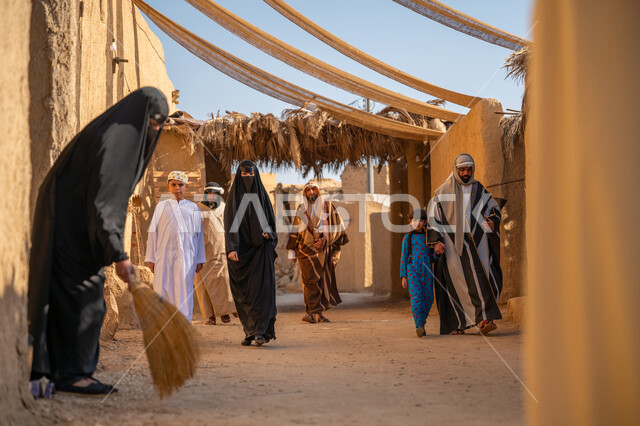 مجموعة من الأشخاص في سوق شعبي قديم، مظلة قماشية ممتدة بين البيوت، يوم بدينا22 فبراير، ذكرى تأسيس الدولة السعودية الأولى1727 م، رجل يرتدي البشت والغترة يمسك بيد ابنته ويتجولان في الحي التاريخي، امرأة عربية خليجية سعودية ترتدي العباءة والنقاب تكنس بمكنسة من القش