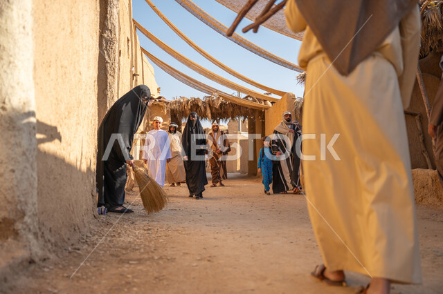 Anniversary of the founding of the first Saudi state 1727 AD, a group of people in a popular market, the day we started, February 22, collecting firewood in the winter season, a close-up photo from the back of a Saudi Arabian Gulf man wearing a traditional thobe and ghutra carrying heavy wood and walking in a historic neighborhood, a cloth stretched between the houses to provide shade