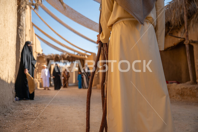 Anniversary of the founding of the first Saudi state 1727 AD, a group of people in a popular market, the day we started, February 22, collecting firewood in the winter season, a close-up photo from the back of a Saudi Arabian Gulf man wearing a traditional thobe and ghutra carrying heavy wood and walking in a historic neighborhood, a cloth stretched between the houses to provide shade