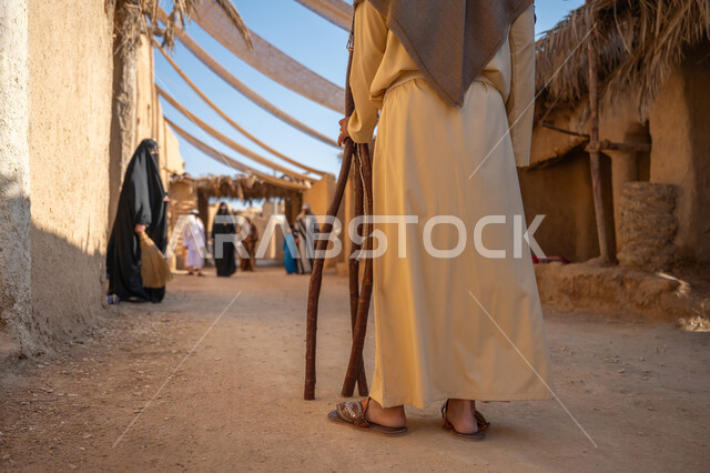 ذكرى تأسيس الدولة السعودية الأولى1727 م، مجموعة من الأشخاص في سوق شعبي، يوم بدينا22 فبراير، تجميع الحطب بموسم الشتاء, صورة مقربة من الخلف لرجل عربي خليجي سعودي يرتدي الثوب التقليدي والغترة يحمل الخشب الثقيل ويمشي في حي تاريخي، قماش ممتد بين البيوت لتوفير الظل