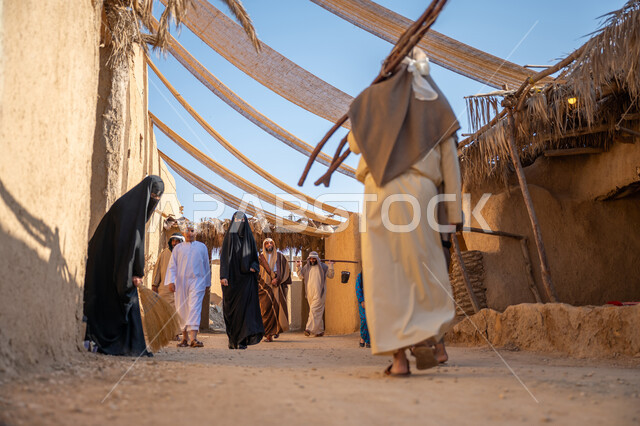 مجموعة من الأشخاص في سوق شعبي، يوم بدينا22 فبراير، ذكرى تأسيس الدولة السعودية الأولى1727 م، تجميع الحطب بموسم الشتاء, صورة مقربة من الخلف لرجل عربي خليجي سعودي يرتدي الثوب التقليدي والغترة يحمل الخشب الثقيل ويمشي في حي تاريخي، قماش ممتد بين البيوت لتوفير الظل