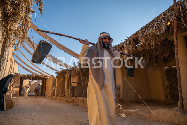 المشي والتجول في سوق شعبي، مهنة شعبية متوارثة، ذكرى تأسيس الدولة السعودية الأولى1727 م، بيت طيني مغطى بسعائف الخوص الجاف، قرية أثرية تاريخية قديمة، يوم بدينا22 فبراير، رجل عربي خليجي سعودي يرتدي الثوب التقليدي والغترة يحمل دلوين بواسطة عصا خشبية طويلة