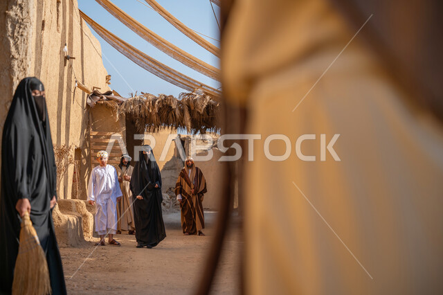 مجموعة من الأشخاص في سوق شعبي، يوم بدينا22 فبراير، ذكرى تأسيس الدولة السعودية الأولى1727 م، تجميع الحطب بموسم الشتاء, صورة مقربة من الخلف لرجل عربي خليجي سعودي يرتدي الثوب التقليدي والغترة يحمل أعواد الخشب في حي تاريخي، قماش ممتد بين البيوت لتوفير الظل