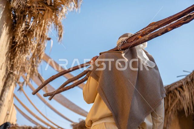 يوم بدينا 22 فبراير، ذكرى تأسيس الدولة السعودية الأولى1727 م، الحياة التقليدية البدوية، استخدام وسائل التدفئه القديمة، تجميع الحطب بموسم الشتاء, صورة مقربة من الخلف لرجل عربي خليجي سعودي يرتدي الثوب التقليدي والغترة يحمل على كتفه الخشب الثقيل ويمشي في حي تاريخي