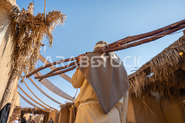 ذكرى تأسيس الدولة السعودية الأولى1727 م، الحياة التقليدية البدوية، استخدام وسائل التدفئه القديمة، يوم بدينا 22 فبراير، تجميع الحطب بموسم الشتاء, صورة مقربة من الخلف لرجل عربي خليجي سعودي يرتدي الثوب التقليدي والغترة يحمل على كتفه الخشب الثقيل ويمشي في حي تاريخي