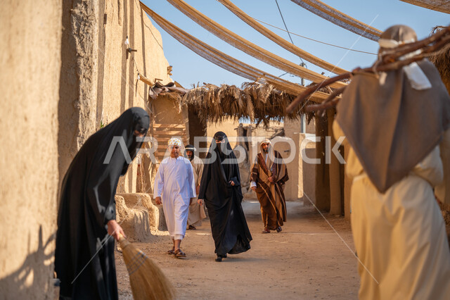 Anniversary of the founding of the first Saudi state 1727 AD, a group of people in a popular market, the day we started, February 22, collecting firewood in the winter season, a close-up photo from the back of a Saudi Arabian Gulf man wearing a traditional thobe and ghutra carrying heavy wood and walking in a historic neighborhood, a cloth stretched between the houses to provide shade