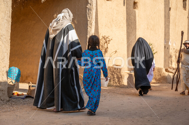 ذكرى تأسيس الدولة السعودية الأولى1727 م، بيت طيني في قرية أثرية قديمة، مجموعة من الأشخاص في سوق شعبي، يوم بدينا22 فبراير، صورة من الخلف لرجل عربي خليجي سعودي يرتدي البشت والغترة يمسك بيد ابنته ويتجولان في الحي التاريخي المبني على الطراز القديم