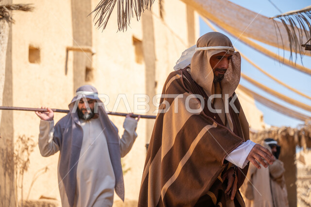 ذكرى تأسيس الدولة السعودية الأولى1727 م، بيت طيني مغطى بسعائف الخوص الجاف، قرية أثرية تاريخية قديمة، التجول في سوق شعبي، يوم بدينا22 فبراير، صورة من الخلف لرجل عربي خليجي سعودي يرتدي الثوب التقليدي والغترة يحمل دلوين بواسطة عصا خشبية طويلة، مهنة شعبية متوارثة