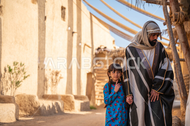 توفير الظل بطريقة قديمة متوارثة، مظلة قماشية ممتدة بين البيوت، مجموعة من الأشخاص في سوق شعبي، يوم بدينا22 فبراير، ذكرى تأسيس الدولة السعودية الأولى1727 م، رجل عربي خليجي سعودي يرتدي البشت والغترة يمسك بيد ابنته ويتجولان في الحي التاريخي المبني على الطراز القديم