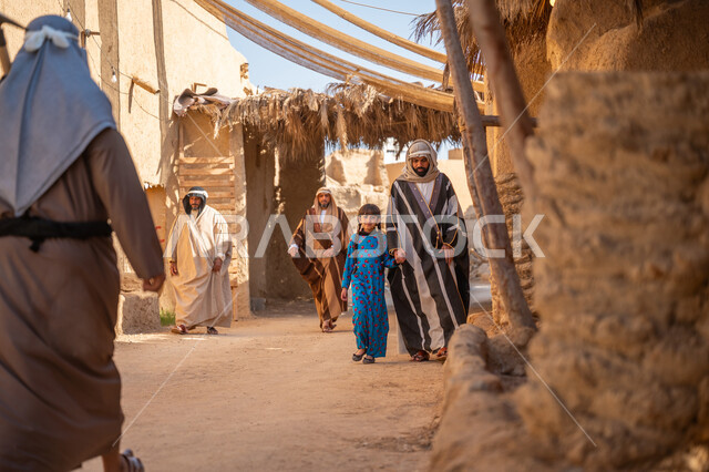 مظلة قماشية ممتدة بين البيوت، توفير الظل بطريقة قديمة متوارثة، مجموعة من الأشخاص في سوق شعبي، يوم بدينا22 فبراير، ذكرى تأسيس الدولة السعودية الأولى1727 م، رجل عربي خليجي سعودي يرتدي البشت والغترة يمسك بيد ابنته ويتجولان في الحي التاريخي المبني على الطراز القديم