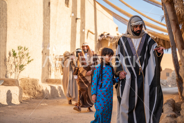 A cloth umbrella stretched between the houses, providing shade in an old inherited way, a group of people in a popular market, on the day of our beginning, February 22, the anniversary of the founding of the first Saudi state 1727 AD, a Saudi Gulf Arab man wearing a bisht and a ghutra holding his daughter's hand as they walk around the historic neighborhood built in the old style