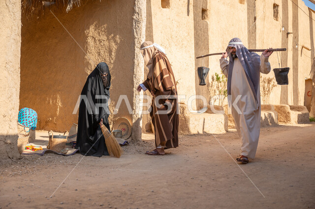 مهنة شعبية متوارثة، ذكرى تأسيس الدولة السعودية الأولى1727 م، بيت طيني مغطى بسعائف الخوص الجاف، قرية أثرية تاريخية قديمة، مجموعة أشخاص يتجولون في سوق شعبي، يوم بدينا22 فبراير، رجل عربي خليجي سعودي يرتدي الثوب التقليدي والغترة يحمل دلوين بواسطة عصا خشبية طويلة