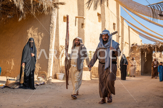Anniversary of the founding of the first Saudi state 1727 AD, a group of people in an ancient popular market, a cloth umbrella extended between the houses, the day we started February 22, two men wearing traditional dress and a ghutra carrying a farming and agricultural tool, wooden branches, walking in the historic neighborhood, a house built in the old style