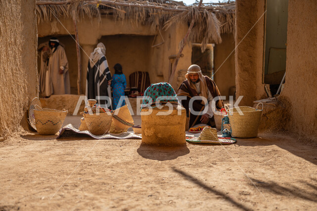 عرض منتج محلي وطني، سوق شعبي تراثي، يوم بدينا 22 فبراير, ذكرى تأسيس الدولة السعودية الأولى1727م، قرية قديمة مبنية من الطوب، سلة من القش مصنوعة بشكل يدوي،  رجل عربي خليجي سعودي يرتدي الغترة والبشت يجلس أمام مشغولات يدوية، منزل طيني قديم مغطى بسعائف الخوص الجاف