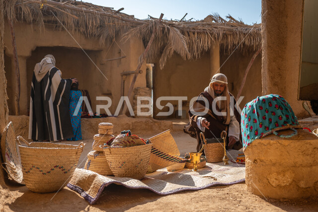 سلة من القش مصنوعة بشكل يدوي، عرض منتج محلي وطني، سوق شعبي تراثي، يوم بدينا 22 فبراير, ذكرى تأسيس الدولة السعودية الأولى1727م، قرية قديمة مبنية من الطوب، رجل عربي خليجي سعودي يرتدي الغترة والبشت يجلس أمام مشغولات يدوية، منزل طيني قديم مغطى بسعائف الخوص الجاف