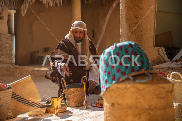 يوم بدينا 22 فبراير, سلة من القش مصنوعة بشكل يدوي، عرض منتج محلي وطني، سوق شعبي تراثي، ذكرى تأسيس الدولة السعودية الأولى1727م، قرية قديمة مبنية من الطوب، رجل عربي خليجي سعودي يرتدي الغترة والبشت يجلس أمام مشغولات يدوية، منزل طيني قديم مغطى بسعائف الخوص الجاف