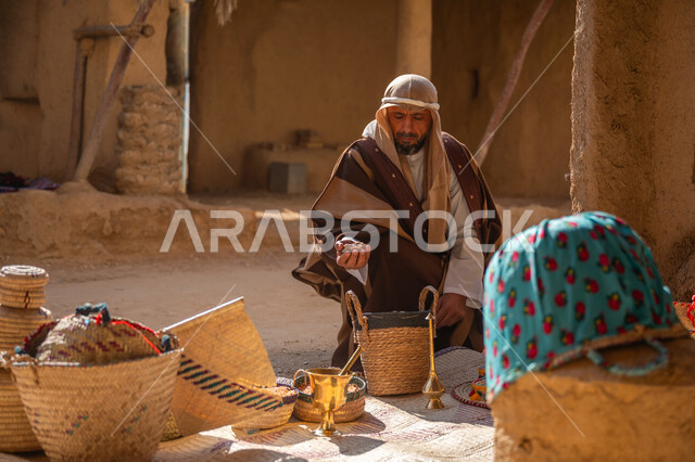سلة من القش مصنوعة بشكل يدوي، عرض منتج محلي وطني، سوق شعبي تراثي، يوم بدينا 22 فبراير, ذكرى تأسيس الدولة السعودية الأولى1727م، قرية قديمة مبنية من الطوب، رجل عربي خليجي سعودي يرتدي الغترة والبشت يجلس أمام مشغولات يدوية، منزل طيني قديم مغطى بسعائف الخوص الجاف