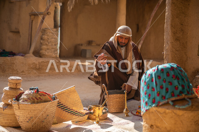 عرض منتج محلي وطني، سوق شعبي تراثي، يوم بدينا 22 فبراير, ذكرى تأسيس الدولة السعودية الأولى1727م، قرية قديمة مبنية من الطوب، سلة من القش مصنوعة بشكل يدوي،  رجل عربي خليجي سعودي يرتدي الغترة والبشت يجلس أمام مشغولات يدوية، منزل طيني قديم مغطى بسعائف الخوص الجاف