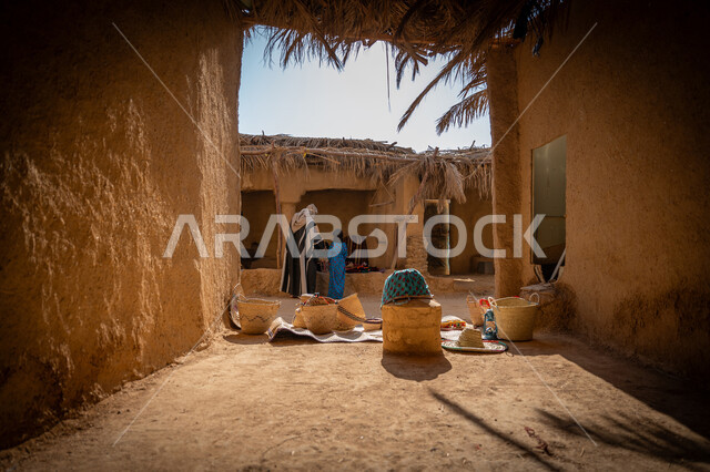 سوق شعبي تراثي، يوم بدينا 22 فبراير, ذكرى تأسيس الدولة السعودية الأولى 1727م، عرض منتج محلي وطني، قرية قديمة مبنية من الطوب، سلة من القش مصنوعة بشكل يدوي، صورة من الخلف لرجل عربي خليجي سعودي يرتدي الغترة والبشت يقف أمام منزل طيني قديم مغطى بسعائف الخوص الجاف