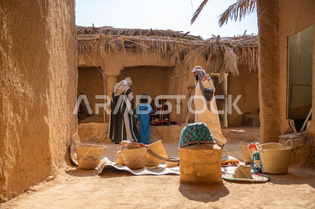 يوم بدينا 22 فبراير, ذكرى تأسيس الدولة السعودية الأولى 1727م، سوق شعبي تراثي، عرض منتج محلي وطني، قرية قديمة مبنية من الطوب، سلة من القش مصنوعة بشكل يدوي، صورة من الخلف لرجل عربي خليجي سعودي يرتدي الغترة والبشت يقف أمام منزل طيني قديم مغطى بسعائف الخوص الجاف