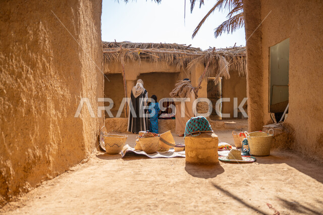 يوم بدينا 22 فبراير, ذكرى تأسيس الدولة السعودية الأولى 1727م، سوق شعبي تراثي، عرض منتج محلي وطني، قرية قديمة مبنية من الطوب، سلة من القش مصنوعة بشكل يدوي، صورة من الخلف لرجل عربي خليجي سعودي يرتدي الغترة والبشت يقف أمام منزل طيني قديم مغطى بسعائف الخوص الجاف