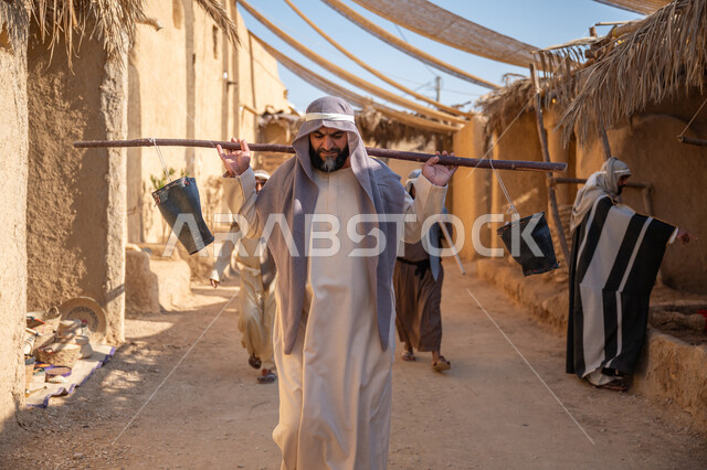 مهنة شعبية متوارثة، ذكرى تأسيس الدولة السعودية الأولى1727 م، بيت طيني مغطى بسعائف الخوص الجاف، قرية أثرية تاريخية قديمة، مجموعة أشخاص يتجولون في سوق شعبي، يوم بدينا22 فبراير، رجل عربي خليجي سعودي يرتدي الثوب التقليدي والغترة يحمل دلوين بواسطة عصا خشبية طويلة