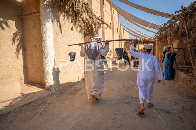 يوم بدينا22 فبراير، مهنة شعبية متوارثة، ذكرى تأسيس الدولة السعودية الأولى1727 م، بيت طيني مغطى بسعائف الخوص الجاف، صبي يمشي في سوق شعبي يقوم بتقديم المساعدة، صورة من الخلف لرجل عربي خليجي سعودي يرتدي الثوب التقليدي والغترة يحمل دلوين بواسطة عصا خشبية طويلة