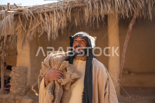 Historical heritage village, the day we started, February 22, the anniversary of the founding of the first Saudi state 1727 AD, a close-up photo of a Saudi Gulf Arab man wearing a bisht and a ghutra standing in front of an old mud house holding a cloth bag and looking somewhere with gestures of pride, the background of the mud house roof covered with dry palm fronds