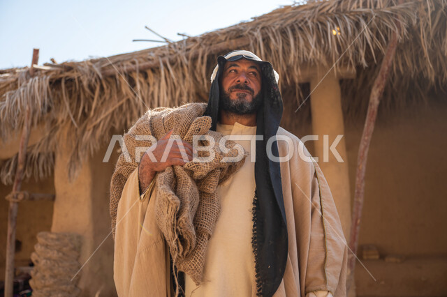 ذكرى تأسيس الدولة السعودية الأولى1727م, قرية تاريخية تراثية، يوم بدينا22 فبراير, صورة مقربة لرجل عربي خليجي سعودي يرتدي البشت والغترة يقف أمام بيت طيني قديم يحمل كيس قماشي وينظر لمكان ما بايماءات الفخر والشموخ، خلفية سقف المنزل الطيني المغطى بسعف النخيل الجاف