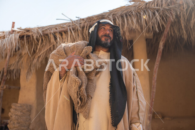 يوم بدينا22 فبراير, ذكرى تأسيس الدولة السعودية الأولى1727م, قرية تاريخية تراثية، صورة مقربة لرجل عربي خليجي سعودي يرتدي البشت والغترة يقف أمام بيت طيني قديم يحمل كيس قماشي وينظر لمكان ما بايماءات الفخر، خلفية سقف المنزل الطيني المغطى بسعف النخيل الجاف