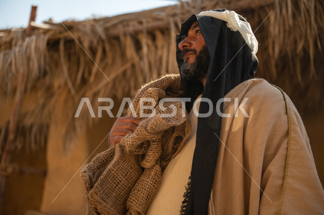 يوم بدينا22 فبراير, ذكرى تأسيس الدولة السعودية الأولى1727م, قرية تاريخية تراثية، صورة مقربة لرجل عربي خليجي سعودي يرتدي البشت والغترة يقف أمام بيت طيني قديم يحمل كيس قماشي وينظر لمكان ما بايماءات الفخر، خلفية سقف المنزل الطيني المغطى بسعف النخيل الجاف