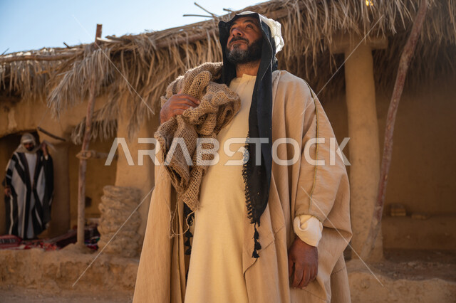 ذكرى تأسيس الدولة السعودية الأولى1727م, قرية تاريخية تراثية، يوم بدينا22 فبراير, رجل عربي خليجي سعودي يرتدي البشت والغترة يقف أمام بيت طيني قديم يحمل كيس قماشي وينظر لمكان ما بايماءات الفخر والشموخ، خلفية سقف المنزل الطيني المغطى بسعف النخيل الجاف