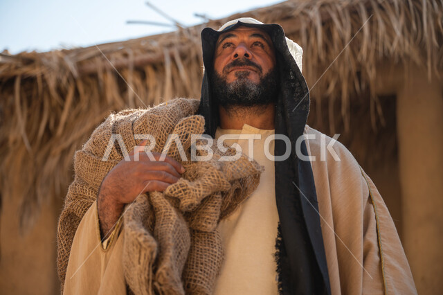 يوم بدينا22 فبراير, ذكرى تأسيس الدولة السعودية الأولى1727م, قرية تاريخية تراثية، صورة مقربة لرجل عربي خليجي سعودي يرتدي البشت والغترة يقف أمام بيت طيني قديم يحمل كيس قماشي وينظر لمكان ما بايماءات الفخر، خلفية سقف المنزل الطيني المغطى بسعف النخيل الجاف