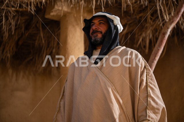 بلدة تاريخية تراثية، يوم بدينا22 فبراير, ذكرى تأسيس الدولة السعودية الأولى1727م, ارتداء الزي الشعبي في مناسبة وطنية, صورة مقربة لرجل عربي خليجي سعودي يرتدي البشت والغترة يقف أمام بيت طيني قديم وينظر للأعلى بايماءات الفخر، خلفية سقف المنزل المغطى بسعف النخيل