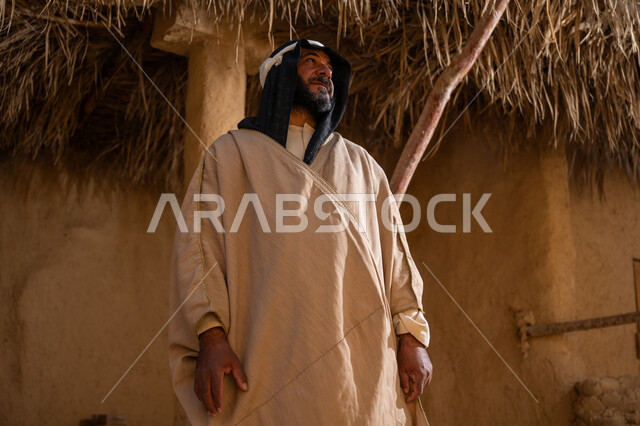 يوم بدينا22 فبراير, بلدة تاريخية تراثية، ذكرى تأسيس الدولة السعودية الأولى1727م, ارتداء الزي الشعبي في مناسبة وطنية, رجل عربي خليجي سعودي يرتدي البشت والغترة يقف أمام بيت طيني قديم وينظر للأعلى بايماءات الفخر، خلفية سقف المنزل المغطى بسعف النخيل