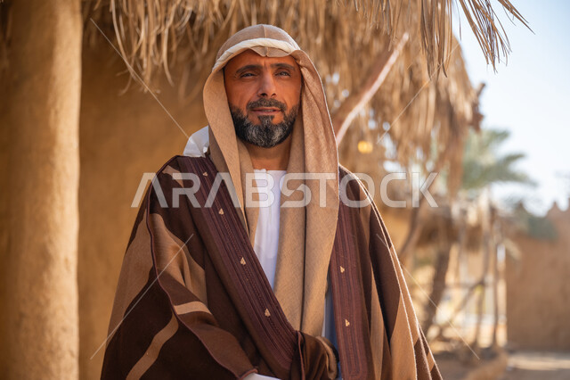 بلدة تاريخية تراثية، يوم بدينا22 فبراير, ذكرى تأسيس الدولة السعودية الأولى1727م, ارتداء الزي الشعبي في مناسبة وطنية, صورة مقربة لرجل عربي خليجي سعودي يرتدي البشت والغترة يقف أمام بيت طيني قديم وينظر للأعلى بايماءات الفخر، خلفية سقف المنزل المغطى بسعف النخيل